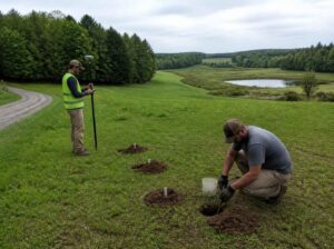 Soil specialist conducting a percolation test while a surveyor records test locations on rural land near a pond.